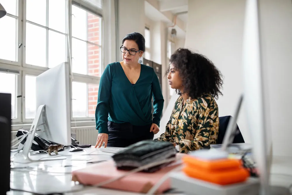 Dua wanita berdiskusi sambil melihat komputer desktop di kantor